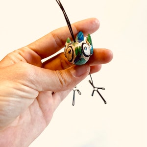 Puede incluir: Una peque&ntilde;a figurita de p&aacute;jaro de cer&aacute;mica pintada en azul, verde y blanco. El p&aacute;jaro tiene ojos grandes y redondos y una cola larga y delgada de alambre.