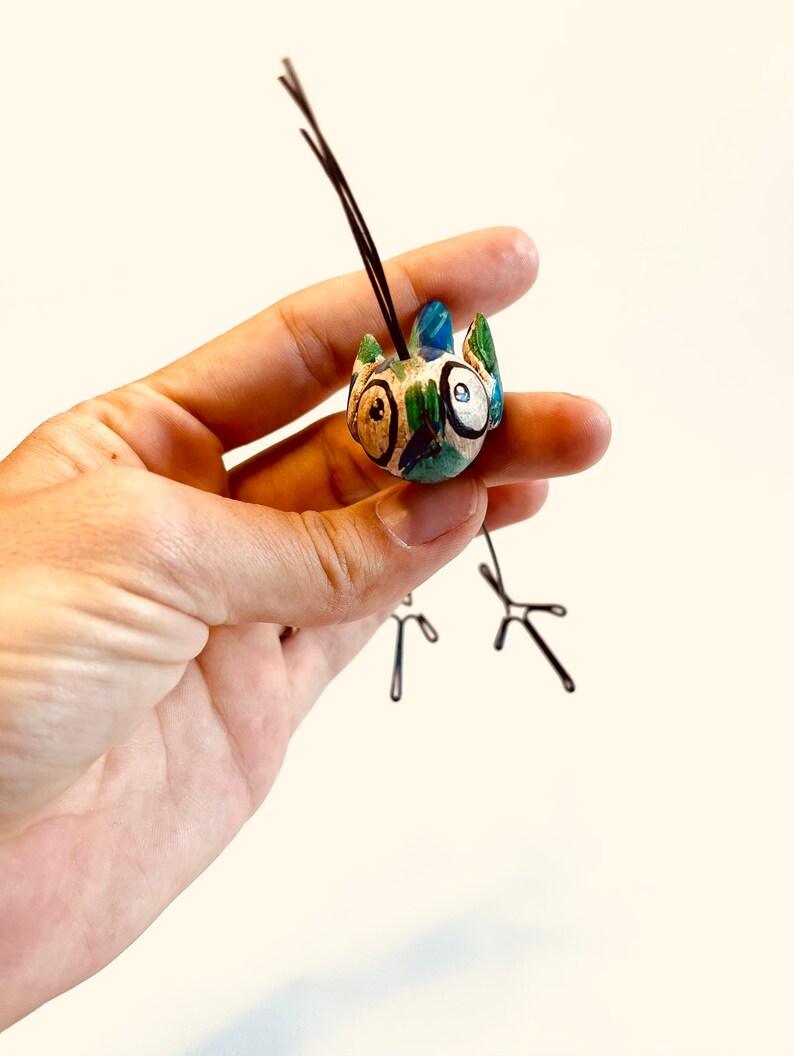 Puede incluir: Una peque&ntilde;a figurita de p&aacute;jaro de cer&aacute;mica pintada en azul, verde y blanco. El p&aacute;jaro tiene ojos grandes y redondos y una cola larga y delgada de alambre.