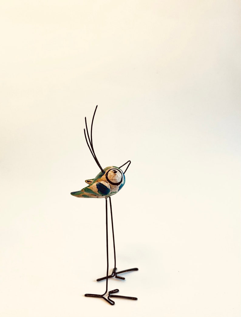 Puede incluir: Una figurita de p&aacute;jaro caprichosa con un cuerpo pintado de azul, verde y blanco, de pie sobre largas y finas patas de alambre negro.