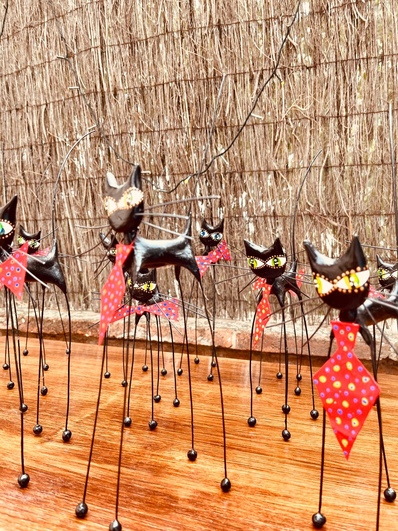 Puede incluir: Un grupo de figuritas de gatos negros con patas largas y delgadas y pajaritas de lunares rojos y blancos. Los gatos est&aacute;n dispuestos en fila sobre una superficie de madera.