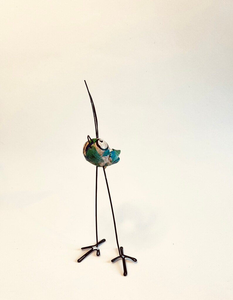 Puede incluir: Una figurita de p&aacute;jaro de cer&aacute;mica caprichosa con un dise&ntilde;o pintado en verde, azul y blanco. El p&aacute;jaro est&aacute; posado sobre largas y delgadas patas de alambre negro.