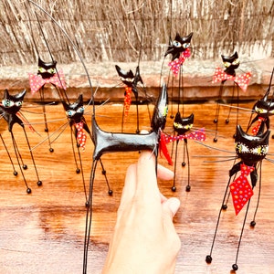 Puede incluir: Una colecci&oacute;n de figuritas de gatos negros con patas largas y delgadas y pajaritas de lunares rojos y blancos. Los gatos est&aacute;n dispuestos sobre una superficie de madera.