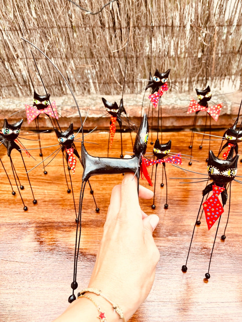 Puede incluir: Una colecci&oacute;n de figuritas de gatos negros con patas largas y delgadas y pajaritas de lunares rojos y blancos. Los gatos est&aacute;n dispuestos sobre una superficie de madera.