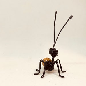 Puede incluir: Escultura de alambre hecha a mano de una hormiga. La hormiga tiene un cuerpo marr&oacute;n con un dise&ntilde;o estampado, patas de alambre negro y dos largas antenas. La escultura est&aacute; sobre un fondo blanco.