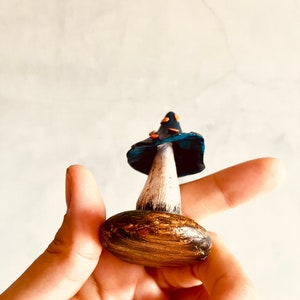 Puede incluir: Una figurita de hongo de cer&aacute;mica pintada a mano con un sombrero azul, un tallo blanco y una base marr&oacute;n. El sombrero tiene manchas naranjas y la base tiene un patr&oacute;n de veta de madera.