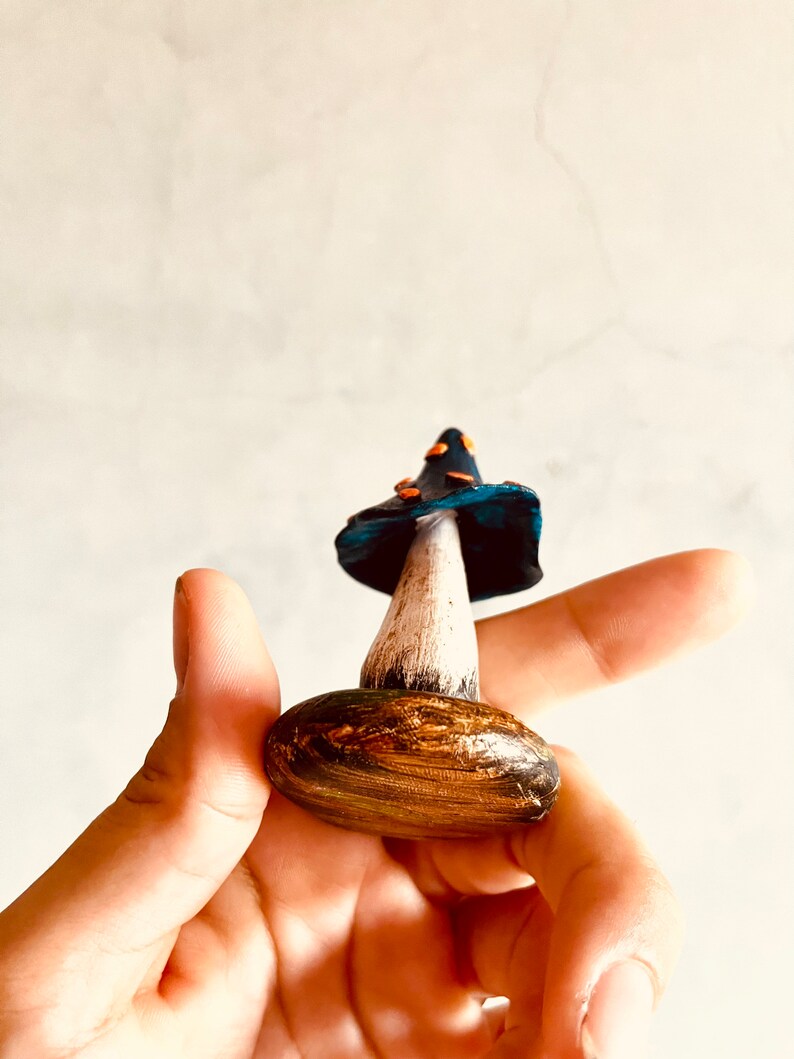 Puede incluir: Una figurita de hongo de cer&aacute;mica pintada a mano con un sombrero azul, un tallo blanco y una base marr&oacute;n. El sombrero tiene manchas naranjas y la base tiene un patr&oacute;n de veta de madera.