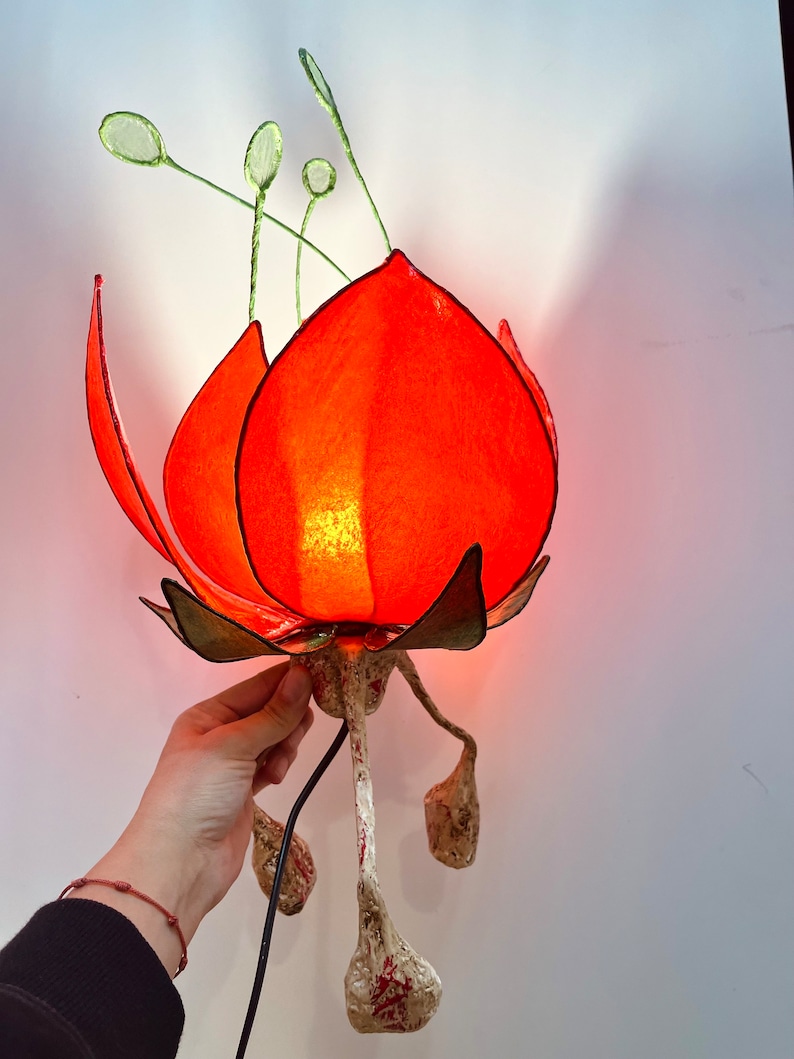 Puede incluir: Una l&aacute;mpara de papel con forma de flor roja con tallos y hojas verdes. La l&aacute;mpara est&aacute; encendida y tiene un cable blanco.