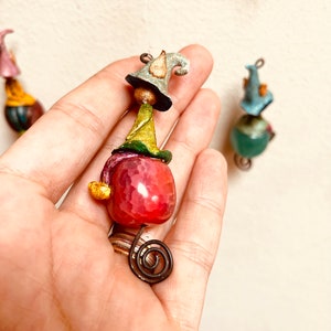 May include: A whimsical gnome ornament with a pink agate stone, green and yellow hat, and a blue and white pointed hat. The gnome is hanging from a thin wire.