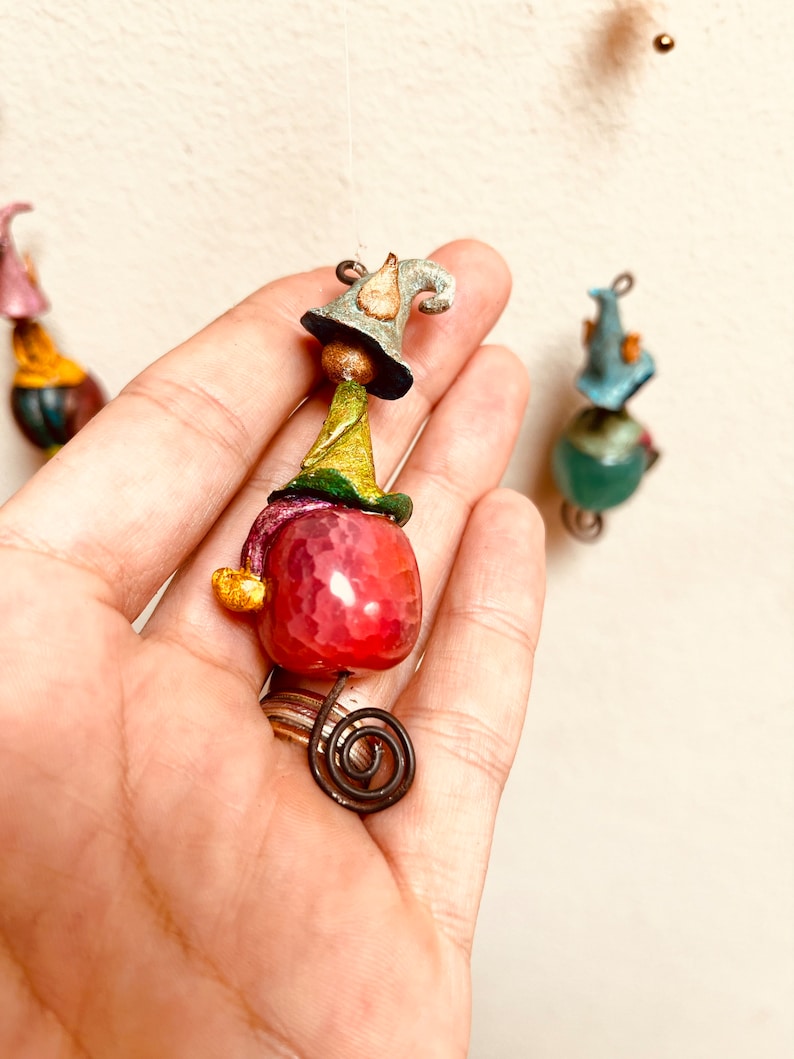 May include: A whimsical gnome ornament with a pink agate stone, green and yellow hat, and a blue and white pointed hat. The gnome is hanging from a thin wire.
