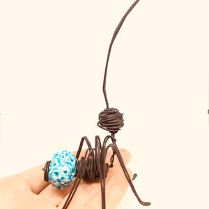 May include: A wire sculpture of an ant with a blue and white patterned bead for a head. The ant is posed with its legs outstretched and its antennae raised.