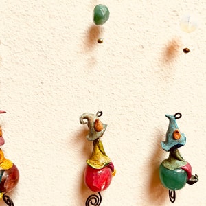 May include: Three whimsical ornaments featuring small, colorful figures with hats, each perched atop a different colored gemstone. The figures are suspended from a string with beads.
