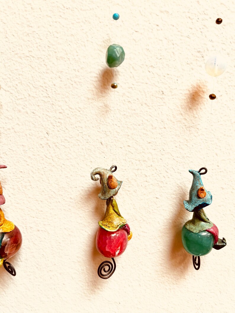 May include: Three whimsical ornaments featuring small, colorful figures with hats, each perched atop a different colored gemstone. The figures are suspended from a string with beads.