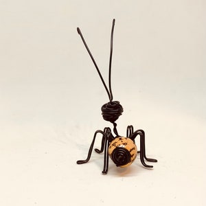 Puede incluir: Una escultura de alambre hecha a mano de una hormiga. La hormiga presenta una cabeza y un cuerpo de alambre enrollado, con seis patas y dos antenas. El cuerpo de la hormiga es de color beige claro con motas marrones oscuras.