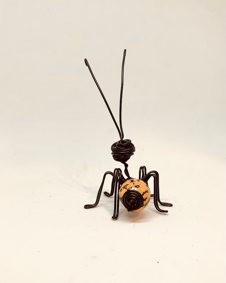Puede incluir: Una escultura de alambre hecha a mano de una hormiga. La hormiga presenta una cabeza y un cuerpo de alambre enrollado, con seis patas y dos antenas. El cuerpo de la hormiga es de color beige claro con motas marrones oscuras.