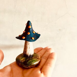 Puede incluir: Una peque&ntilde;a figura de hongo azul y naranja con tallo blanco y base verde. El hongo est&aacute; pintado a mano y tiene una est&eacute;tica caprichosa de cuento de hadas.