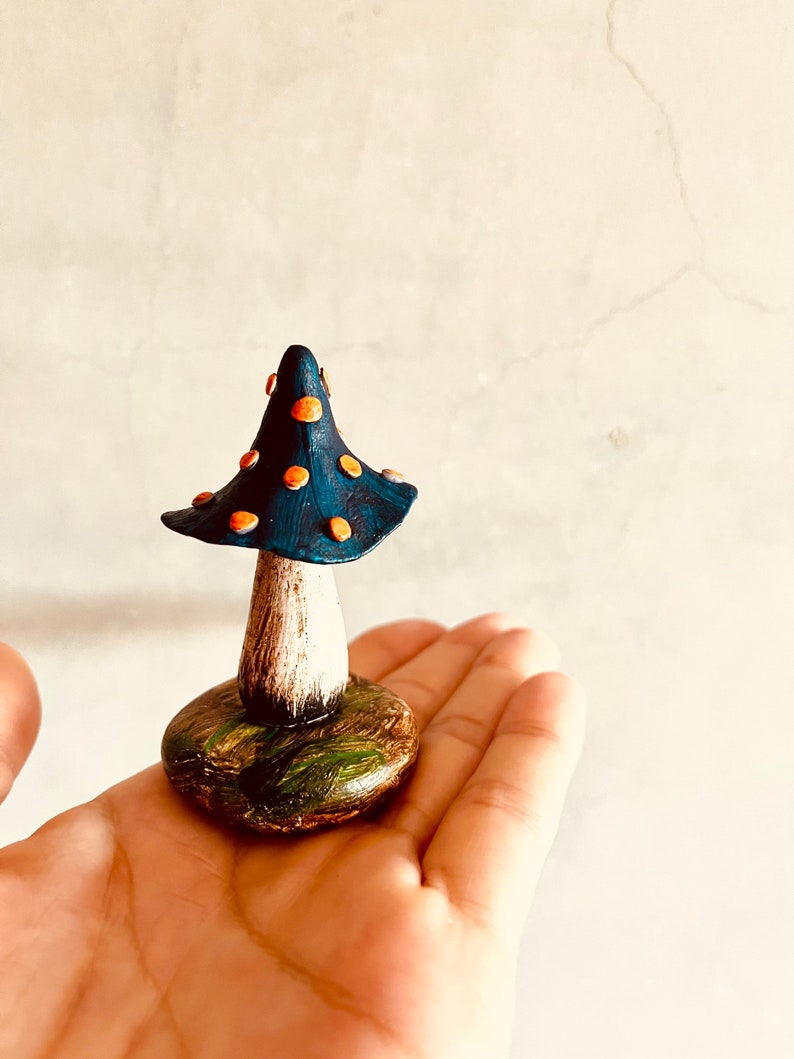 Puede incluir: Una peque&ntilde;a figura de hongo azul y naranja con tallo blanco y base verde. El hongo est&aacute; pintado a mano y tiene una est&eacute;tica caprichosa de cuento de hadas.