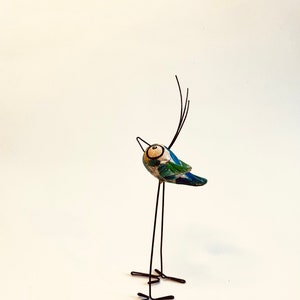 Puede incluir: Una figurita de p&aacute;jaro azul y verde caprichosa con un cuerpo y patas de alambre negro. El p&aacute;jaro tiene un ojo blanco grande y un pico largo y delgado.