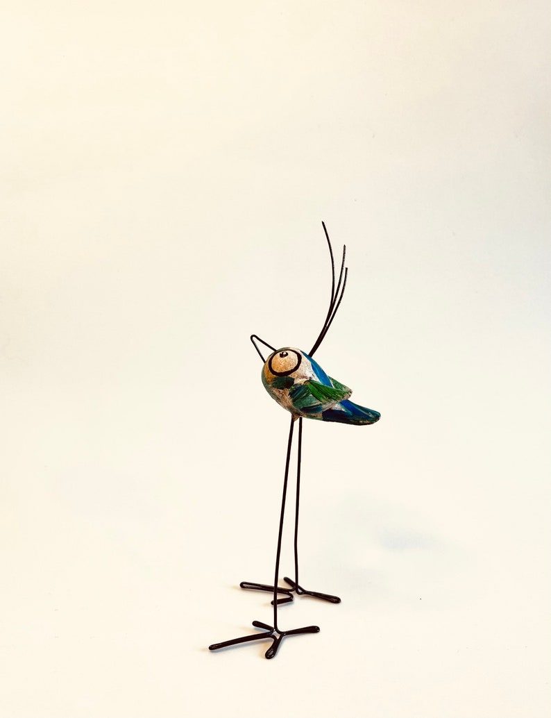 Puede incluir: Una figurita de p&aacute;jaro azul y verde caprichosa con un cuerpo y patas de alambre negro. El p&aacute;jaro tiene un ojo blanco grande y un pico largo y delgado.