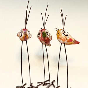 Puede incluir: Tres figuritas de p&aacute;jaros caprichosas con patas largas y delgadas y detalles pintados. Cada p&aacute;jaro tiene un esquema de color diferente y presenta ojos grandes y redondos. Los p&aacute;jaros est&aacute;n hechos de una combinaci&oacute;n de alambre y material pintado.
