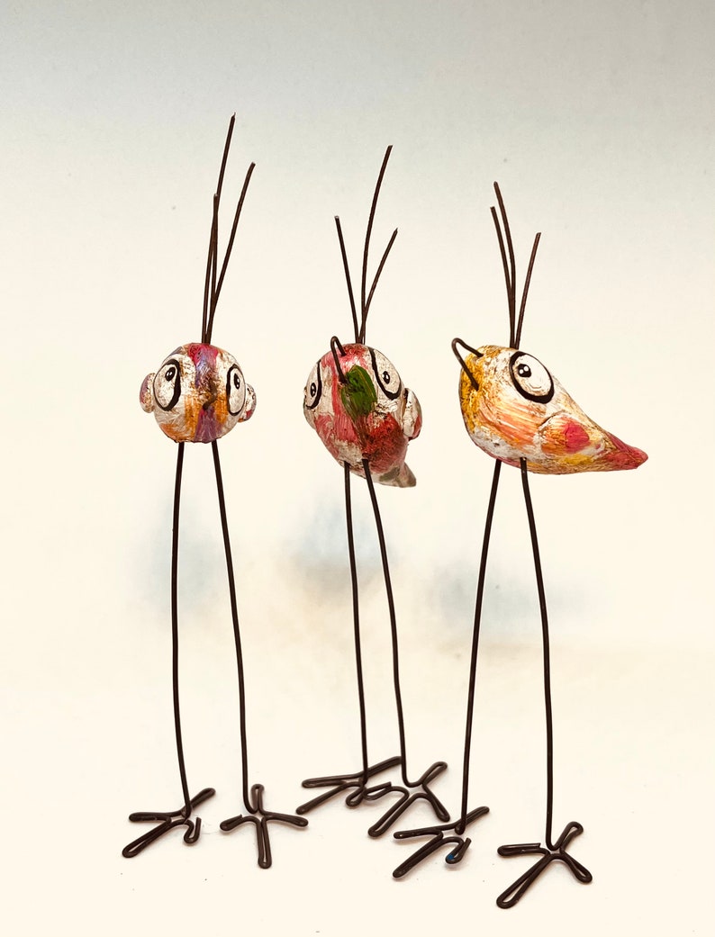 Puede incluir: Tres figuritas de p&aacute;jaros caprichosas con patas largas y delgadas y detalles pintados. Cada p&aacute;jaro tiene un esquema de color diferente y presenta ojos grandes y redondos. Los p&aacute;jaros est&aacute;n hechos de una combinaci&oacute;n de alambre y material pintado.