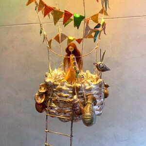 Może przedstawiać: Kapryśna, ręcznie robiona rzeźba balonu na gorące powietrze. Kosz jest teksturowany i zawiera małą figurkę z długimi, rudymi włosami. Kolorowe chorągiewki i lampki choinkowe zdobią balon, do kt&oacute;rego przymocowana jest mała figurka ptaka. Wisząca drabina.