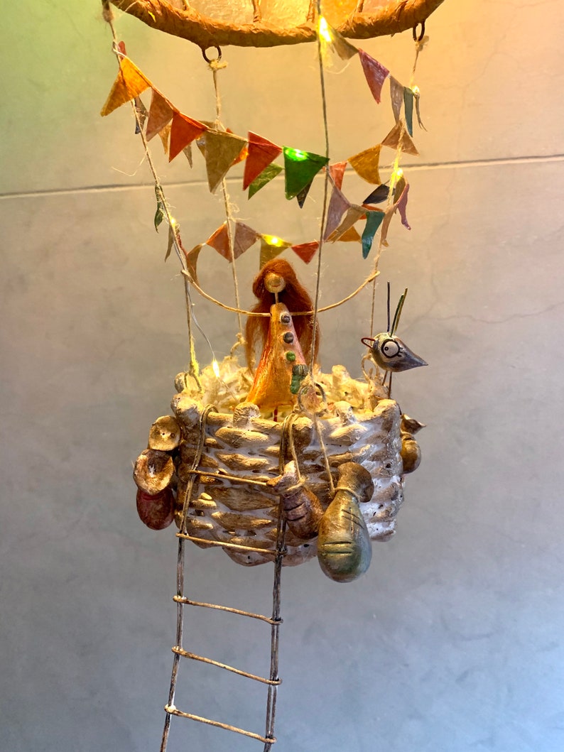 Może przedstawiać: Kapryśna, ręcznie robiona rzeźba balonu na gorące powietrze. Kosz jest teksturowany i zawiera małą figurkę z długimi, rudymi włosami. Kolorowe chorągiewki i lampki choinkowe zdobią balon, do kt&oacute;rego przymocowana jest mała figurka ptaka. Wisząca drabina.