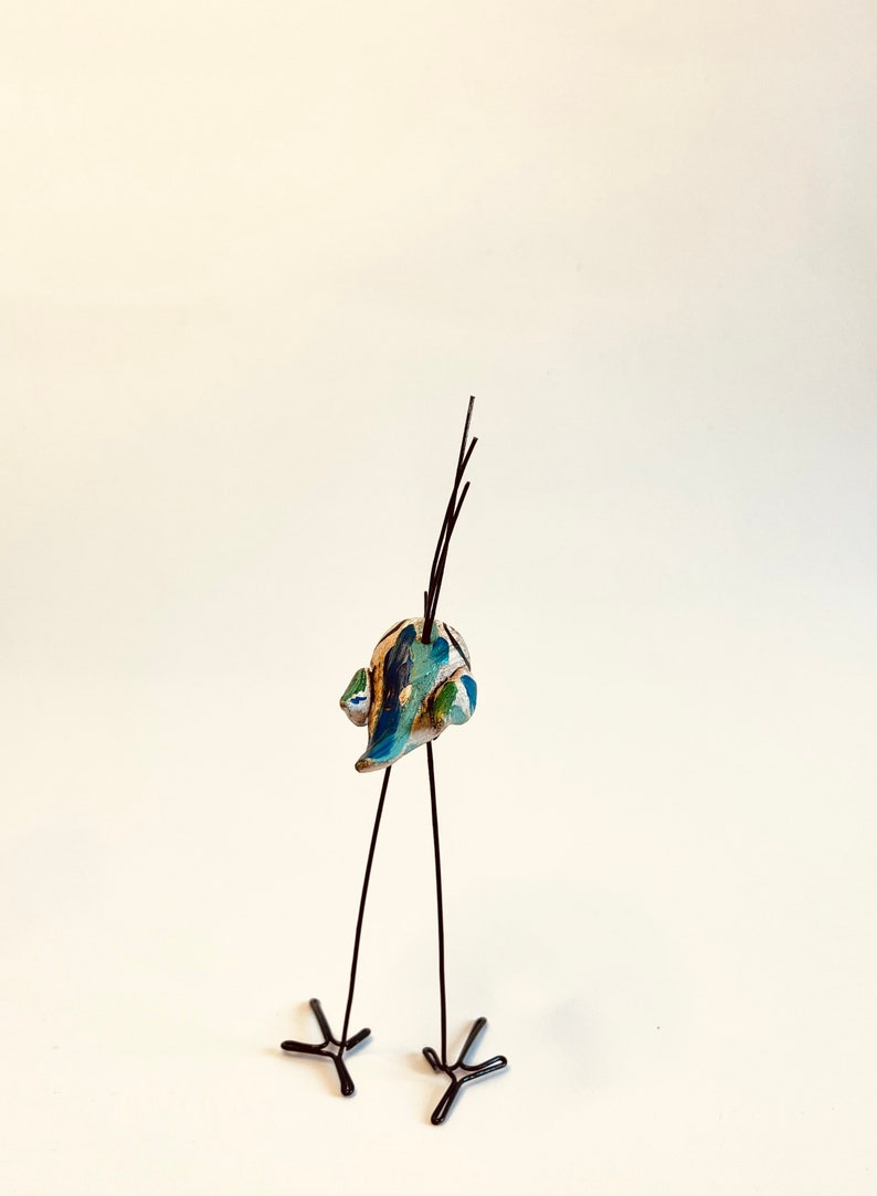 Puede incluir: Una peque&ntilde;a escultura de p&aacute;jaro de cer&aacute;mica con un cuerpo estampado en azul, verde y blanco. El p&aacute;jaro tiene patas largas y delgadas de alambre negro y una cola larga y delgada de alambre negro.