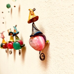 May include: A pink, green, and red mobile with a whimsical design. Each piece features a small, colorful figure with a hat and a unique shape. The mobile is suspended from a white ceiling.