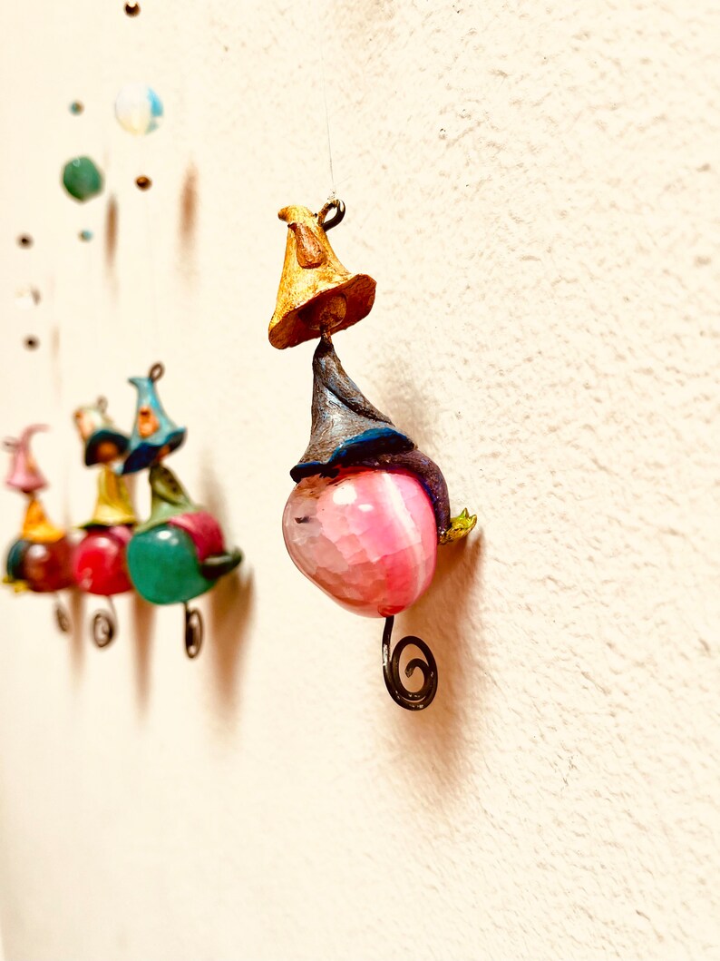 May include: A pink, green, and red mobile with a whimsical design. Each piece features a small, colorful figure with a hat and a unique shape. The mobile is suspended from a white ceiling.