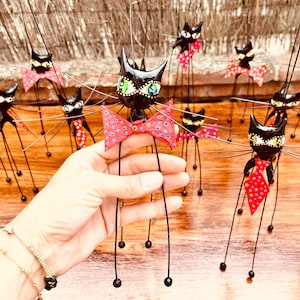 Puede incluir: Un grupo de figuritas de gatos negros con pajaritas de lunares rojos y blancos. Los gatos tienen patas largas y delgadas y est&aacute;n hechos de un material negro. Los gatos est&aacute;n de pie sobre una superficie de madera.