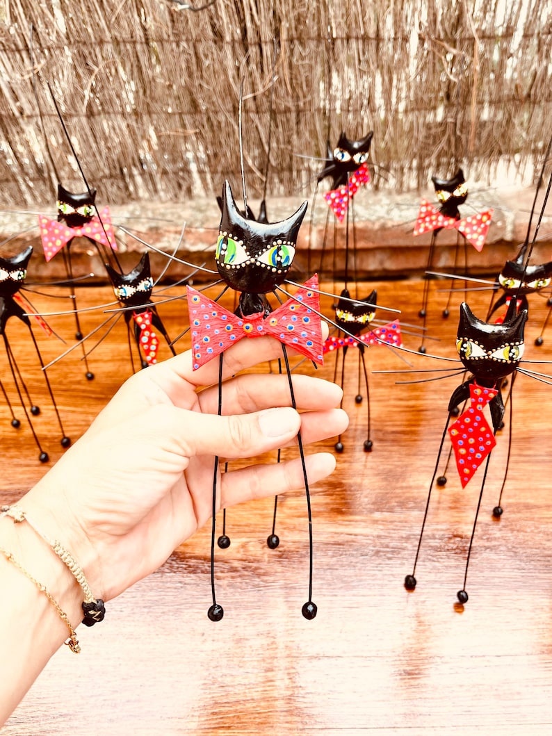 Puede incluir: Un grupo de figuritas de gatos negros con pajaritas de lunares rojos y blancos. Los gatos tienen patas largas y delgadas y est&aacute;n hechos de un material negro. Los gatos est&aacute;n de pie sobre una superficie de madera.