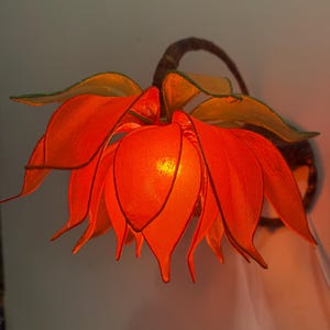 May include: A decorative wall sconce shaped like a large orange flower with green-edged petals. The lamp has a warm, inviting glow, and is mounted on a dark brown, curved stem.