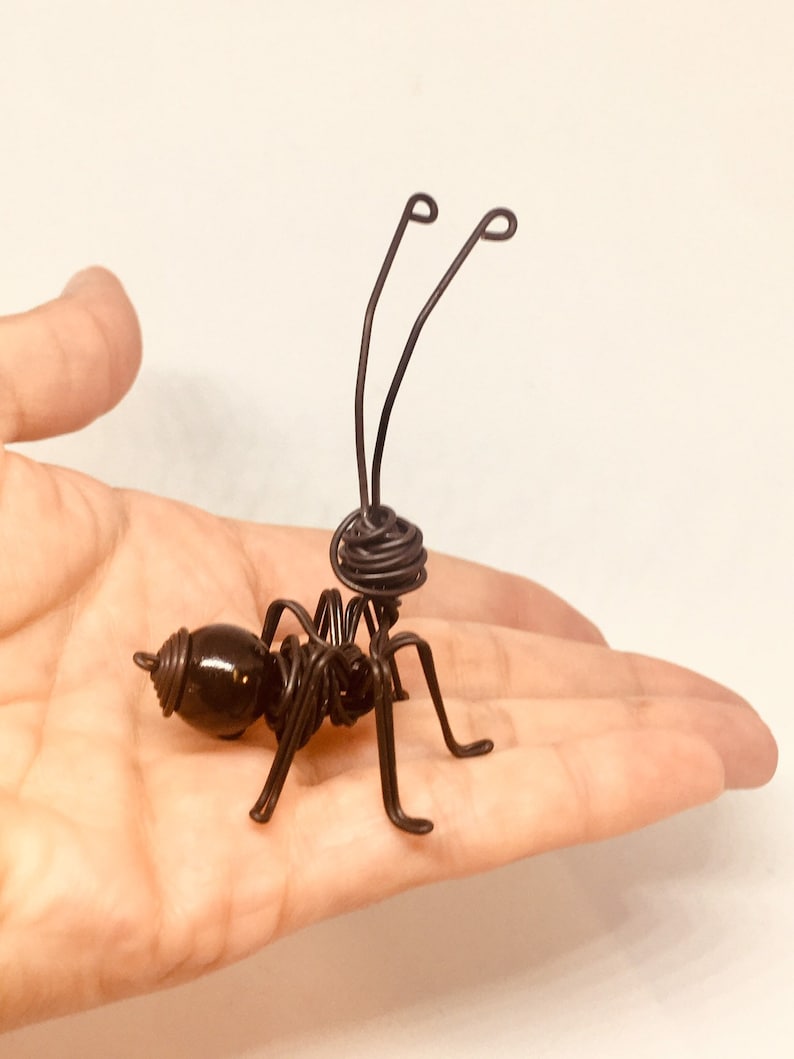 K&ouml;nnte beinhalten: Eine schwarze Drahtskulptur einer Ameise mit einer schwarzen Perle als Kopf. Die Ameise ist mit ausgestreckten Beinen und erhobenen F&uuml;hlern dargestellt.