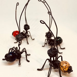 Puede incluir: Cuatro hormigas artesanales de alambre con cuerpos de cuentas de vidrio en rojo, verde, azul y beige. Las hormigas tienen cuerpos de alambre enrollado, seis patas de alambre y antenas largas y curvas. Esculturas &uacute;nicas.