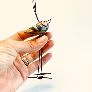 Puede incluir: Un p&aacute;jaro de cer&aacute;mica caprichoso con detalles pintados en azul y verde, posado sobre un soporte de alambre negro. El p&aacute;jaro tiene un pico largo y delgado y un cuerpo redondo.