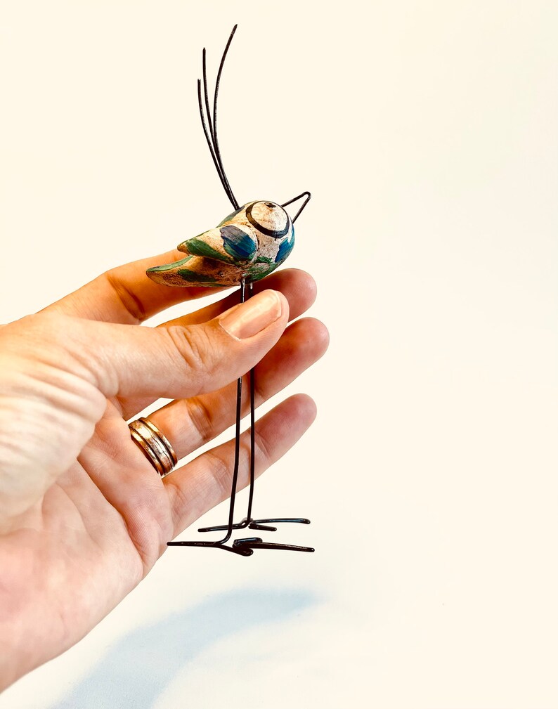 Puede incluir: Un p&aacute;jaro de cer&aacute;mica caprichoso con detalles pintados en azul y verde, posado sobre un soporte de alambre negro. El p&aacute;jaro tiene un pico largo y delgado y un cuerpo redondo.