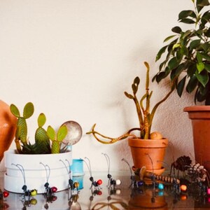 Puede incluir: Un bodeg&oacute;n con varias plantas en macetas, incluyendo cactus y una planta de hojas verdes. Hormigas decorativas de metal con cuerpos de cuentas de colores est&aacute;n dispuestas frente a las plantas. Tambi&eacute;n se ve una maceta grande de terracota y una maceta de cer&aacute;mica blanca.