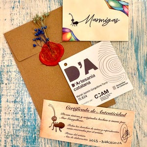Puede incluir: Un conjunto de tarjetas artesanales y un sobre sobre una superficie de madera azul y blanca desgastada. Las tarjetas presentan ilustraciones de hormigas y texto, incluyendo "Marmigas" y un certificado de autenticidad. Un sello de cera rojo y flores secas adornan el sobre.