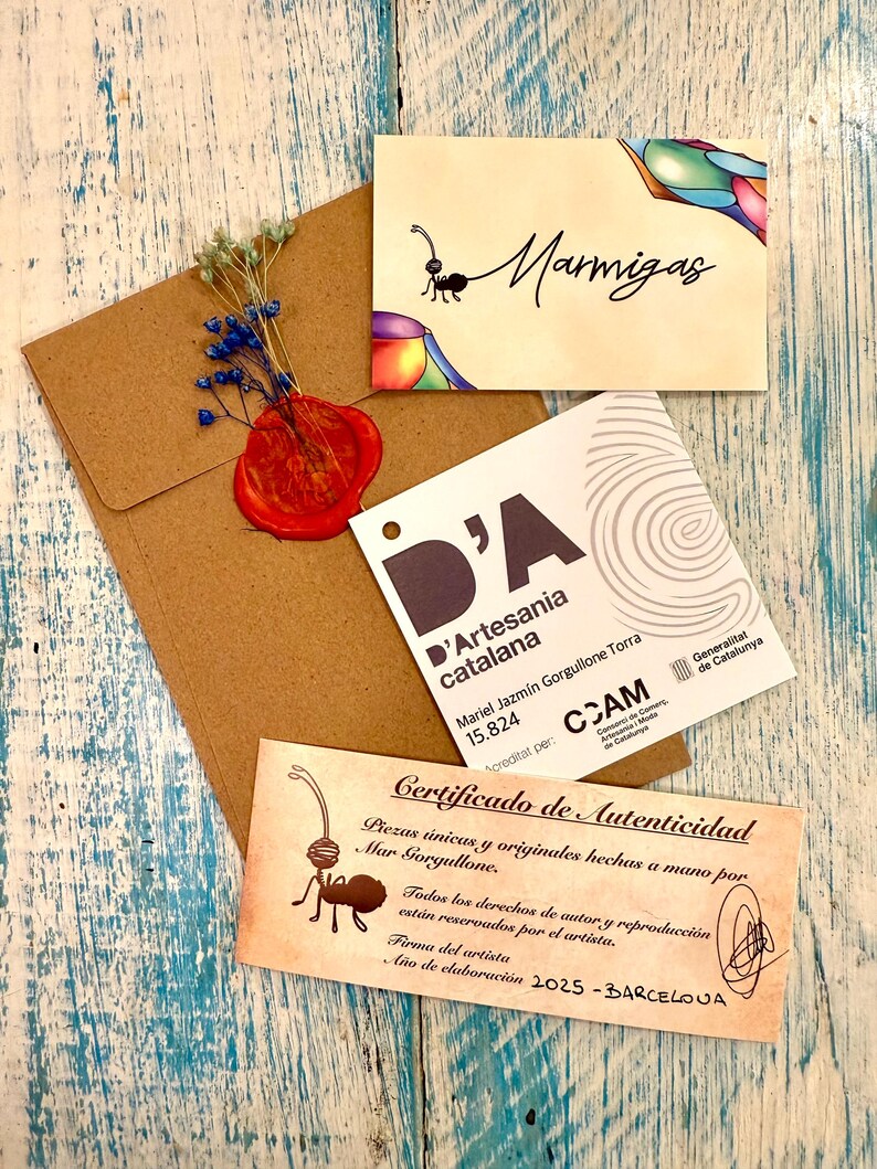 Puede incluir: Un conjunto de tarjetas artesanales y un sobre sobre una superficie de madera azul y blanca desgastada. Las tarjetas presentan ilustraciones de hormigas y texto, incluyendo "Marmigas" y un certificado de autenticidad. Un sello de cera rojo y flores secas adornan el sobre.