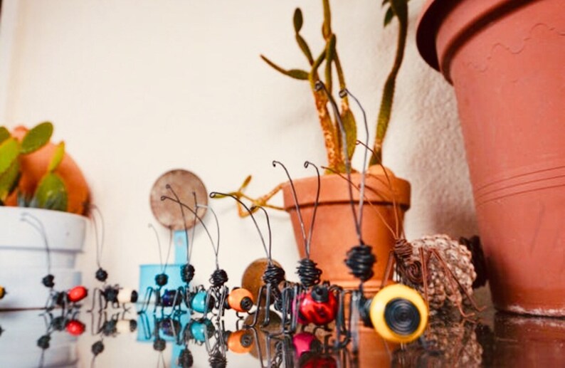 Puede incluir: Una colecci&oacute;n de esculturas de insectos de alambre caprichosas con detalles coloridos. Los insectos tienen cuerpos negros, patas de alambre y antenas. Algunos tienen detalles rojos, azules y amarillos. Una maceta de terracota con una planta est&aacute; en el fondo.