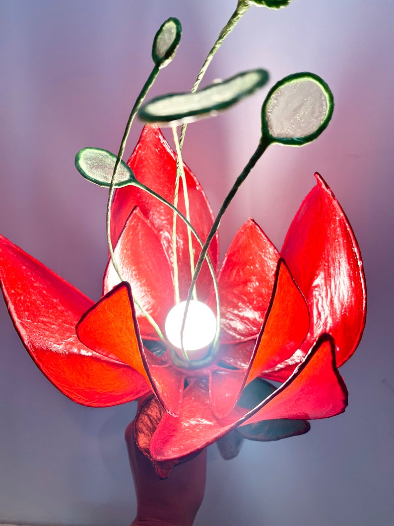 Puede incluir: Una l&aacute;mpara de papel con forma de flor roja con tallos y hojas verdes. La l&aacute;mpara tiene una bombilla blanca en el centro.