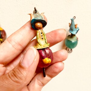 May include: A whimsical figurine made of paper mache, featuring a small person wearing a blue hat, a green shirt, and a red skirt. The figurine is attached to a wire loop.