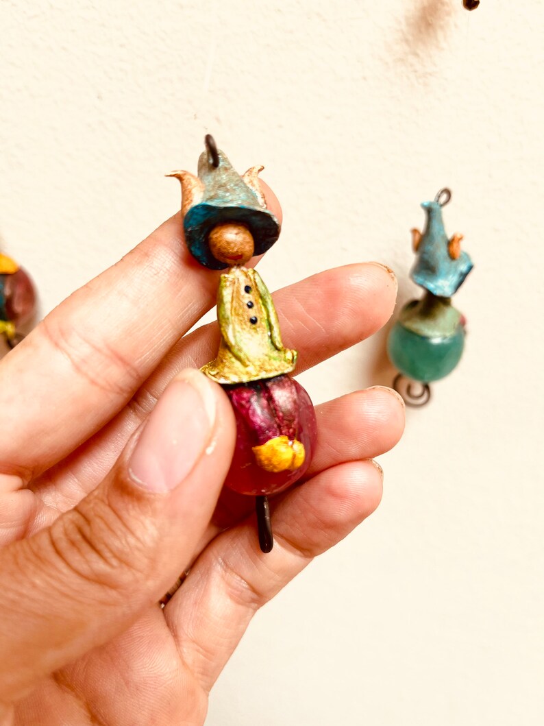 May include: A whimsical figurine made of paper mache, featuring a small person wearing a blue hat, a green shirt, and a red skirt. The figurine is attached to a wire loop.