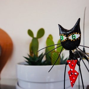 Puede incluir: Figurilla de gato negro con ojos verdes y una pajarita roja con lunares blancos. La figurilla est&aacute; hecha de metal y tiene un cuerpo largo y delgado. El gato est&aacute; de pie sobre una mesa con una planta en maceta en el fondo.