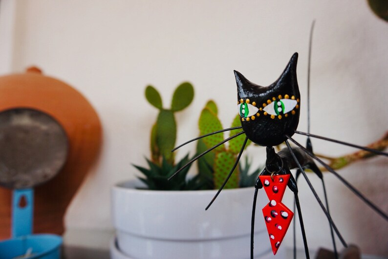 Puede incluir: Figurilla de gato negro con ojos verdes y una pajarita roja con lunares blancos. La figurilla est&aacute; hecha de metal y tiene un cuerpo largo y delgado. El gato est&aacute; de pie sobre una mesa con una planta en maceta en el fondo.