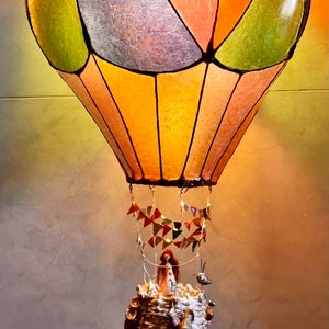 Może przedstawiać: Kapryśna lampa w kształcie balonu na ogrzane powietrze z kolorowym, segmentowym kloszem w kolorze pomarańczowym, zielonym i fioletowym. Kosz posiada drabinę, miniaturowe figurki i małego ptaszka. Ozdobne proporczyki wiszą poniżej.