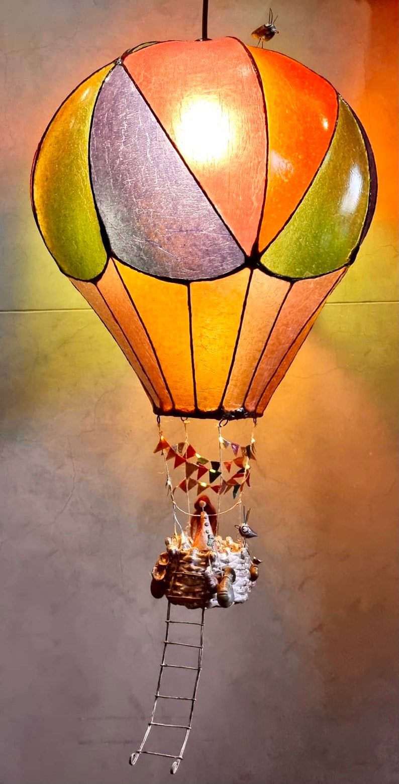 Może przedstawiać: Kapryśna lampa w kształcie balonu na ogrzane powietrze z kolorowym, segmentowym kloszem w kolorze pomarańczowym, zielonym i fioletowym. Kosz posiada drabinę, miniaturowe figurki i małego ptaszka. Ozdobne proporczyki wiszą poniżej.