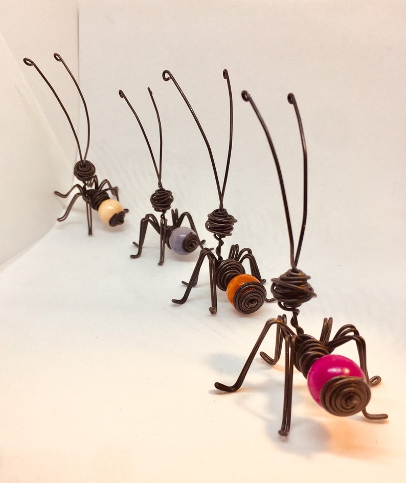 Puede incluir: Una fila de hormigas de arte de alambre hechas a mano. Cada hormiga tiene un cuerpo, patas y antenas de alambre marr&oacute;n oscuro. Las hormigas tienen coloridos abd&oacute;menes de cuentas en tonos de rosa, naranja, lavanda y crema. Las hormigas est&aacute;n dispuestas en diagonal.