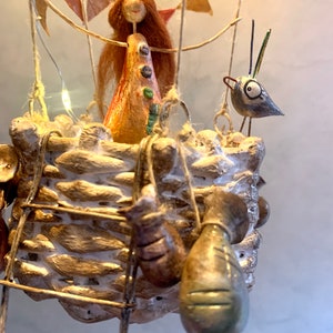 Może przedstawiać: Ręcznie wykonana rzeźba balonu na gorące powietrze. Kosz jest tkany, ze szczeblem i wiszącymi workami. Postać z rudymi włosami i ptak znajdują się w balonie. Kolorowe chorągiewki i lampki choinkowe dopełniają świąteczny, dekoracyjny design.