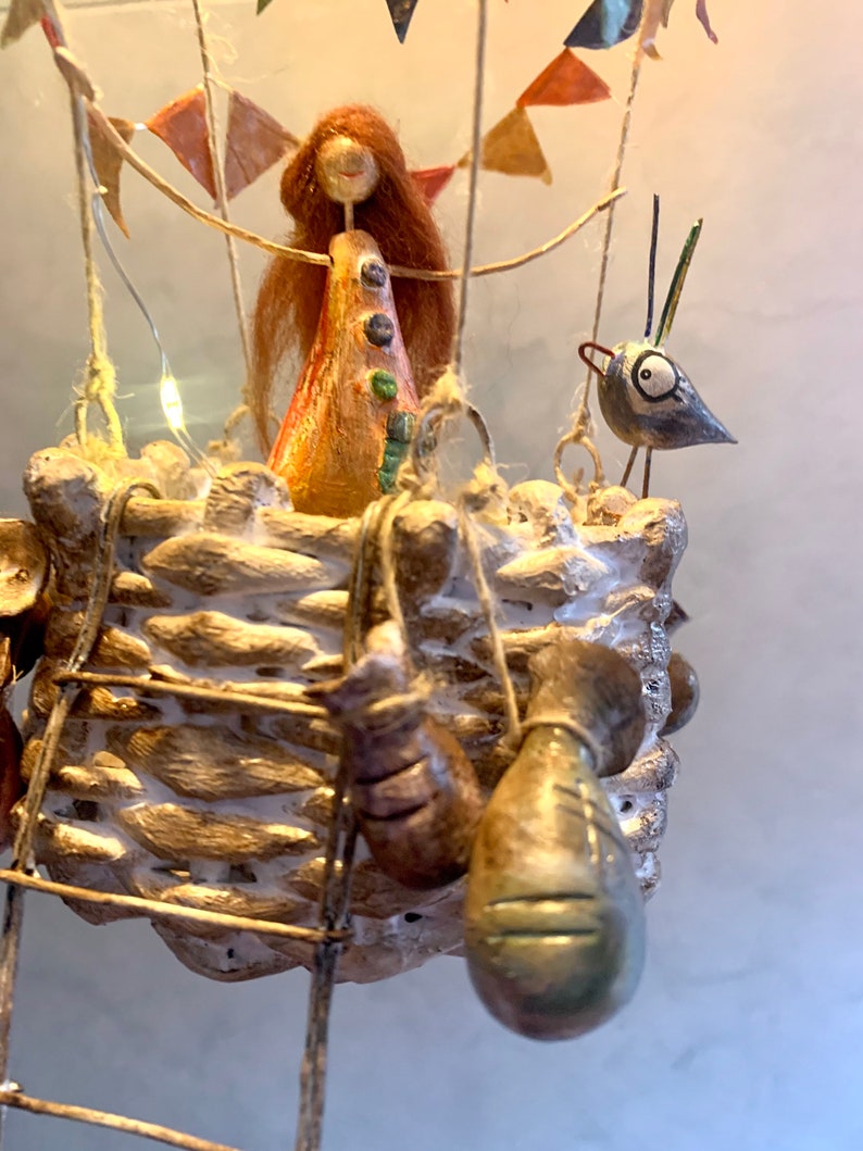 Może przedstawiać: Ręcznie wykonana rzeźba balonu na gorące powietrze. Kosz jest tkany, ze szczeblem i wiszącymi workami. Postać z rudymi włosami i ptak znajdują się w balonie. Kolorowe chorągiewki i lampki choinkowe dopełniają świąteczny, dekoracyjny design.